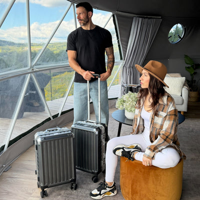 Couple in a glamping dome with two gray aluminum-frame LUGGEX suitcases, a carry-on and a checked size, with a scenic valley view behind them.#color_charcoal